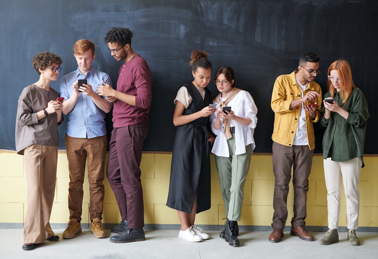 Young diverse adults using smartphones while standing against a blackboard, showcasing technology and connection.